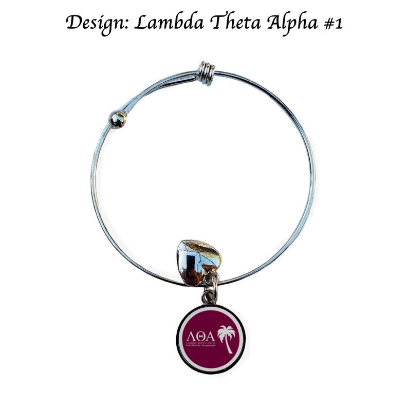 Puede incluir: Una pulsera de brazalete plateada con un dije redondo. El dije es un c&iacute;rculo rosa oscuro con texto blanco y un gr&aacute;fico de palmera. El texto "Lambda Theta Alpha #1" est&aacute; encima de la pulsera.