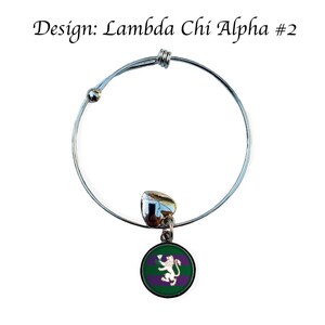 Puede incluir: Un brazalete extensible de plata con un dije que presenta un c&iacute;rculo verde y morado con un le&oacute;n blanco y una flor blanca. El dije est&aacute; unido a un dije de coraz&oacute;n plateado. El brazalete est&aacute; etiquetado como "Dise&ntilde;o: Lambda Chi Alpha #2".