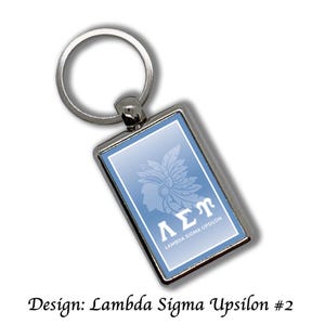 Op de afbeelding: Zilveren rechthoekige sleutelhanger met een lichtblauwe achtergrond. De sleutelhanger heeft een witte afbeelding van een profiel met een gevederde hoofdtooi en de Griekse letters ΛΣΠ. De tekst "LAMBDA SIGMA UPSILON" is ook aanwezig.