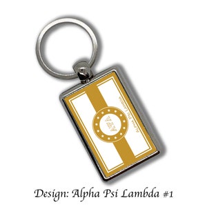 Può includere: Un portachiavi d'argento con un'etichetta rettangolare che presenta un design bianco e oro. L'etichetta ha un cerchio dorato con le lettere "ΑΨΑ" in bianco e una fascia dorata con il testo "ALPHA PSI LAMBDA" in bianco.