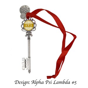 May include: A silver key-shaped Christmas ornament with a gold and white patterned circle featuring the letters "ΑΨΛ" and the text "Alpha Psi Lambda". The ornament is attached to a red ribbon.