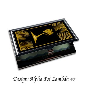Peut inclure: Un porte-cartes de visite noir et or avec un motif de palmier et les lettres "A&Psi;&Lambda;" sur le devant. Le porte-cartes est ouvert pour r&eacute;v&eacute;ler les emplacements pour les cartes.