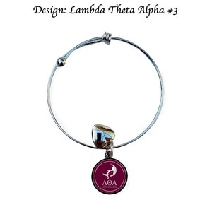 Puede incluir: Una pulsera de brazalete plateada con un dije redondo de color granate. El dije presenta un gr&aacute;fico blanco y las letras griegas &Lambda;&Theta;&Alpha;. La pulsera tiene un peque&ntilde;o acento en forma de coraz&oacute;n y el texto "Design: Lambda Theta Alpha #3".