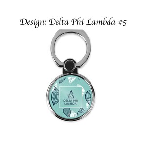 Op de afbeelding: Een zilveren telefoonringhouder met een rond, turquoise ontwerp met witte en marineblauwe bladerpatronen. Het midden heeft een witte vierkant met de tekst "DELTA PHI LAMBDA". De tekst "Design: Delta Phi Lambda #5" staat erboven.