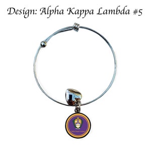 Puede incluir: Un brazalete extensible plateado con un dije que presenta un logotipo de Alpha Kappa Lambda morado y dorado.