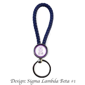 Puede incluir: Un llavero de cuero trenzado azul marino con un anillo de metal plateado y un emblema redondo. El emblema presenta un borde morado y un centro blanco con las letras "ZΛB" y un escudo. El texto "Design: Sigma Lambda Beta #1" está en la parte inferior.
