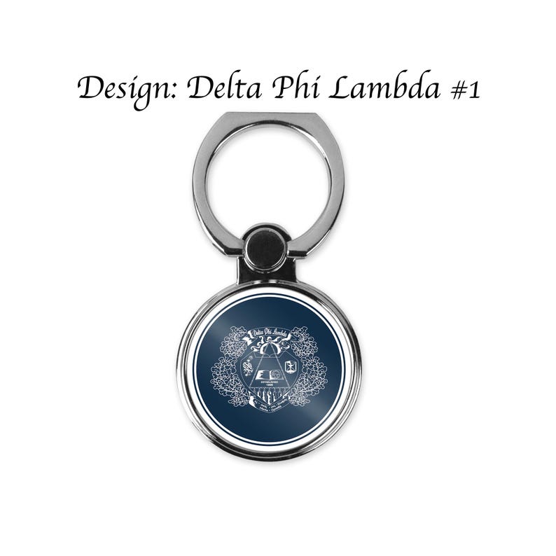 Op de afbeelding: Een zilveren telefoonringhouder met een cirkelvormig ontwerp. Het ontwerp heeft een donkerblauwe achtergrond met een wit embleem en de tekst "Delta Phi Lambda #1". De ring is bevestigd aan een draaibare basis.