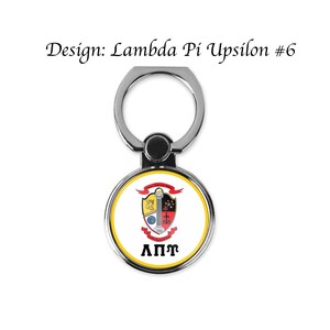 Op de afbeelding: Een zilveren telefoonringhouder met een rond, wit, rood en geel embleem met een schild met een kolom, een wereldbol, een ster en een kruis. De tekst "&Lambda;&Pi;&Gamma;" is onder het embleem gedrukt.