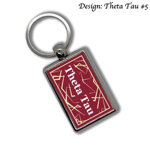 May include: A rectangular keychain with a silver-colored metal frame. The maroon insert features the words "Theta Tau" in white, with gold geometric lines. The design is labeled "Design: Theta Tau #5".