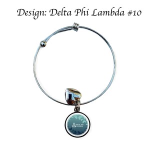 K&ouml;nnte beinhalten: Silbernes, ausziehbares Armband mit einem runden Anh&auml;nger, der ein gr&uuml;nes und wei&szlig;es Tarnmuster mit dem Text "Delta Phi Lambda" in Wei&szlig; zeigt.