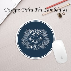 Może przedstawiać: Okrągła, ciemnoniebieska podkładka pod mysz z białym wzorem herbu przedstawiającego książkę, klepsydrę i inne symbole. Herb jest otoczony wieńcem kwiat&oacute;w, a widoczny jest napis "Delta Phi Lambda Established 1968".