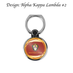 K&ouml;nnte beinhalten: Ein silberner Handy-Ringhalter mit einem runden, roten, gelben und goldenen Design. Das Design zeigt ein Wappen mit einem lila und wei&szlig;en Symbol und dem Text "Alpha Kappa Lambda".