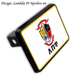 May include: Black trailer hitch cover with a white rectangular face. The face features a colorful crest with a pillar, scales, and a globe, with the Greek letters &Lambda;&Pi;&Upsilon; and the text "Lambda Pi Upsilon #6". The border is yellow.