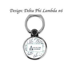 Op de afbeelding: Een zilveren telefoonringhouder met een ronde, patroonvormige voorkant. De voorkant heeft een bloemenontwerp in blauw- en groentinten, met de tekst "DELTA PHI LAMBDA" in een rechthoekig kader. De tekst "Design: Delta Phi Lambda #6" staat boven de ring.