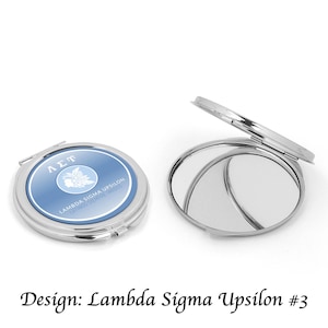 Op de afbeelding: Twee zilveren compacte spiegels. Eén is gesloten en toont een blauw-wit ontwerp met tekst. De andere is open en onthult twee spiegelende oppervlakken. De tekst op de gesloten compact luidt "Lambda Sigma Upsilon".