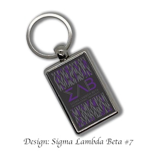 Può includere: Un portachiavi argentato con un'etichetta rettangolare nera e grigia. L'etichetta presenta un design a motivi viola e grigi con le lettere "ΣΛΒ" e il testo "Sigma Lambda Beta International Fraternity".