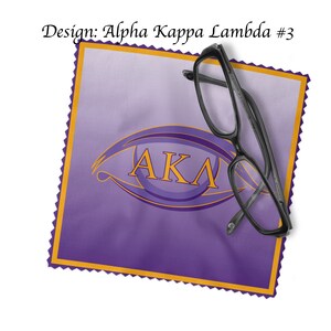 K&ouml;nnte beinhalten: Lilafarbener Mikrofaser-Reinigungstuch mit goldfarbenem Rand und einem goldenen Augenmotiv mit den Buchstaben "AKA" darin. Ein Paar schwarze Brillen liegt auf dem Tuch.