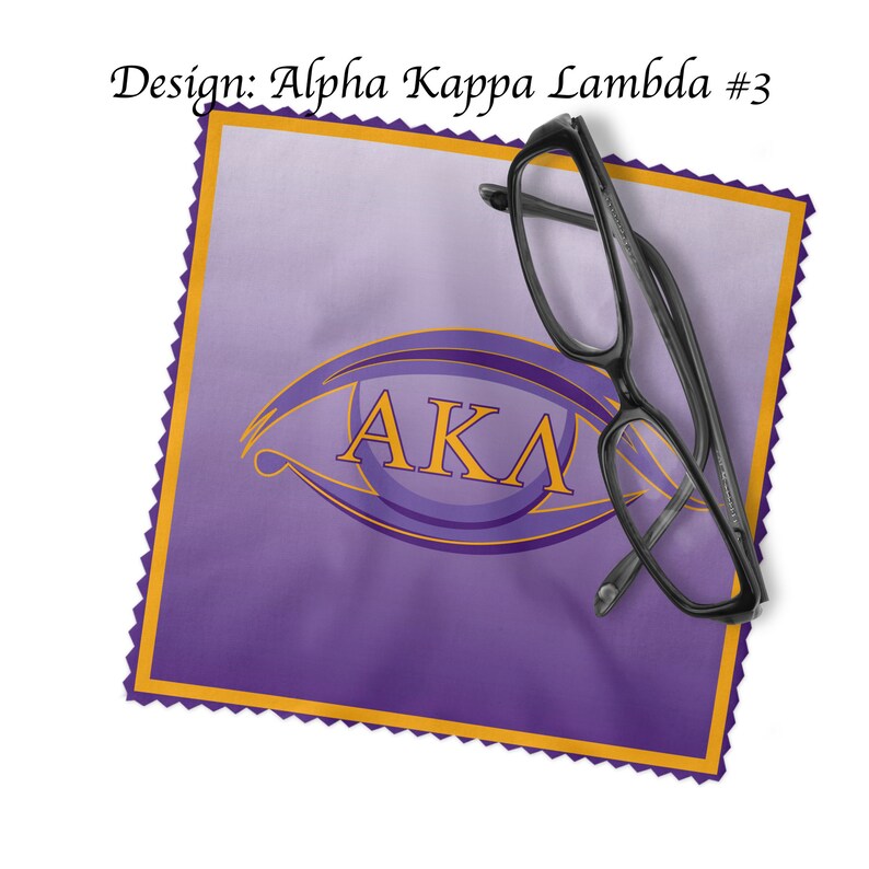 K&ouml;nnte beinhalten: Lilafarbener Mikrofaser-Reinigungstuch mit goldfarbenem Rand und einem goldenen Augenmotiv mit den Buchstaben "AKA" darin. Ein Paar schwarze Brillen liegt auf dem Tuch.