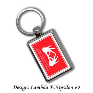 Op de afbeelding: Een rechthoekige zilveren sleutelhanger met een rode achtergrond met een witte afbeelding en tekst. De tekst luidt "LAMBDA PI UPSILON SORORITY LATINAS PODEROSAS UNIDAS, INC." en "Design: Lambda Pi Upsilon #2."