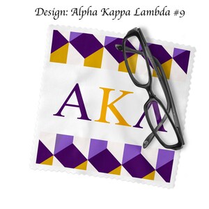 K&ouml;nnte beinhalten: Ein wei&szlig;es Mikrofaser-Reinigungstuch mit einem geometrischen Muster in Lila und Gold und den Buchstaben "AKA" in Gold. Ein Paar schwarzer Brillen liegt auf dem Tuch.