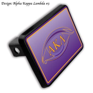 Può includere: Un copri gancio di traino nero con un design viola e oro. Il design presenta le lettere "AKA" all'interno di una forma di occhio stilizzata. Il testo "Design: Alpha Kappa Lambda #3" si trova nell'angolo in alto a sinistra.