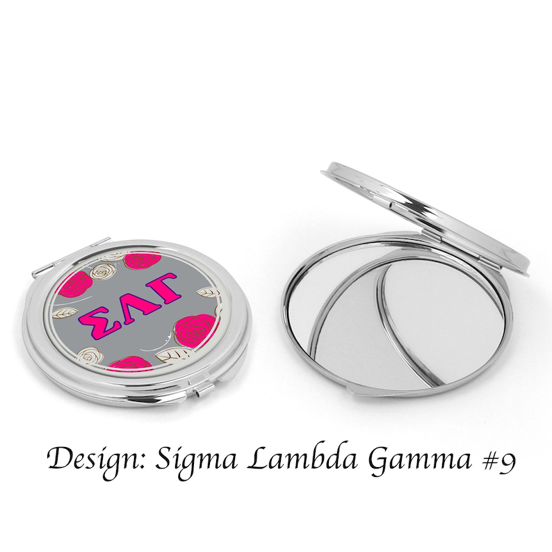Op de afbeelding: Een zilveren compact spiegel met een grijze achtergrond en roze rozen. De spiegel heeft de letters "&Sigma;&Lambda;&Gamma;" in roze op de voorkant.