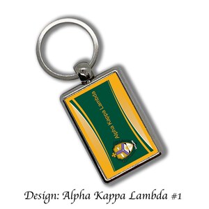 May include: A silver keychain with a rectangular tag featuring a gold and green design. The tag has the text "Alpha Kappa Lambda" and a crest with purple and gold accents.