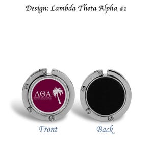 May include: A silver-colored, round, foldable bag hook with a burgundy circle on the front featuring a white palm tree and the text "ΛΘΑ Lambda Theta Alpha, Inc.". The back of the hook is black.