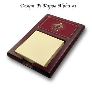 Peut inclure: Un porte-bloc-notes de bureau en bois avec un blason Pi Kappa Alpha rouge et or sur le dessus. Le porte-bloc-notes a une fente pour un bloc-notes et un fond rouge avec des accents dorés.