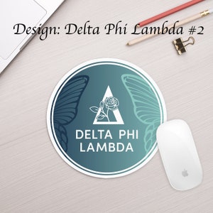 Może przedstawiać: Okrągła podkładka pod mysz w kolorze turkusowym i niebieskim z białym obramowaniem. W środku znajduje się biały tr&oacute;jkąt z r&oacute;żą w środku i tekstem "DELTA PHI LAMBDA". Po obu stronach tr&oacute;jkąta znajdują się dwa białe skrzydła motyla.