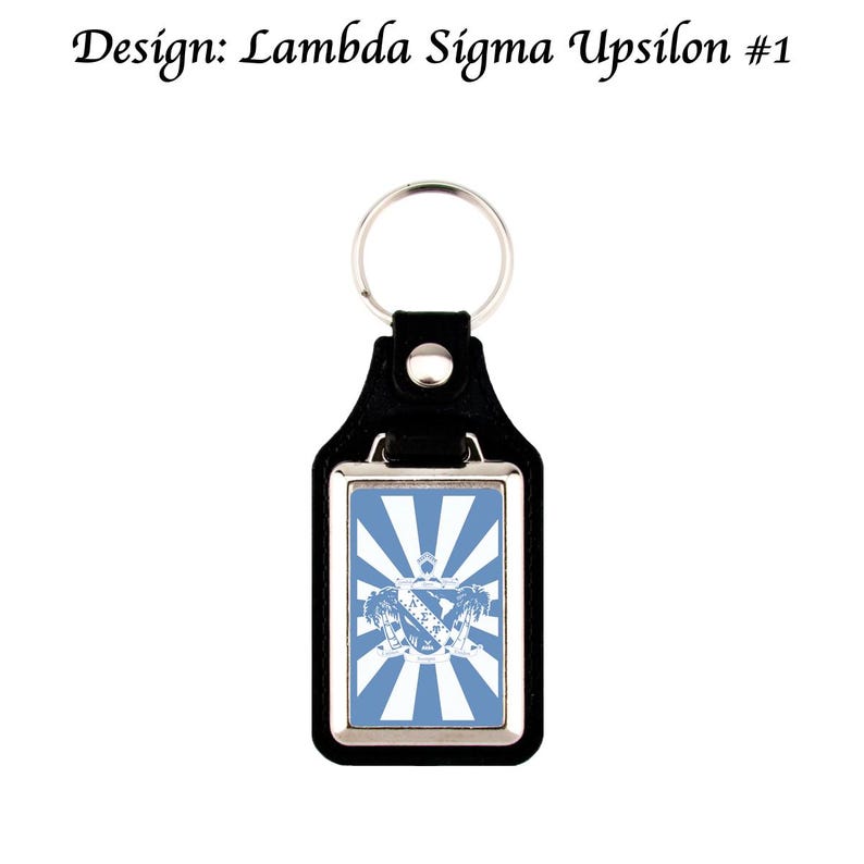 May include: Black leather keychain with a silver metal frame. The rectangular insert has a blue and white design with a crest and the text "Design: Lambda Sigma Upsilon #1". Includes a metal keyring.