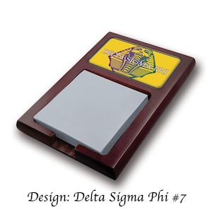 May include: A wooden desk notepad holder with a recessed area for a stack of grey notepaper. The top section has a yellow background with the words "DELTA SIGMA PHI" and a graphic in purple and green.