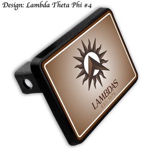 Puede incluir: Cubierta negra para enganche de remolque con un diseño marrón y blanco. El diseño presenta un logotipo de sol y flecha con el texto "LAMBDAS EST. 1975". El texto "Design: Lambda Theta Phi #4" está en la esquina superior izquierda.