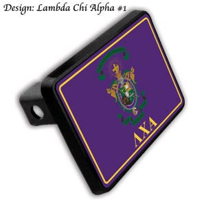 May include: A black trailer hitch cover featuring a purple background with a gold border. The cover displays a crest with green, gold, and white accents, and the Greek letters ΛΧΑ in gold. The text "Design: Lambda Chi Alpha #1" is at the top.
