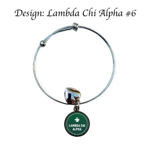 Puede incluir: Un brazalete extensible de plata con un dije verde que dice "Lambda Chi Alpha" en letras blancas. El dije tambi&eacute;n tiene una cruz blanca.