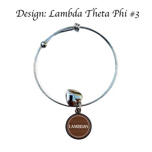 Op de afbeelding: Een zilveren uittrekbaar armband met een bruine bedel met de tekst "LAMBDAS" in wit. De bedel heeft een gepatroneerd ontwerp.