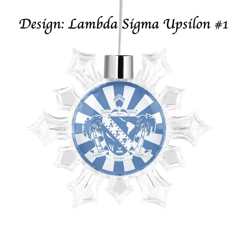 May include: A blue and white snowflake ornament with a Lambda Sigma Upsilon crest. The crest features a shield with the Greek letters Lambda, Sigma, and Upsilon, along with a palm tree and the words "Latinos Siempre Unidos".