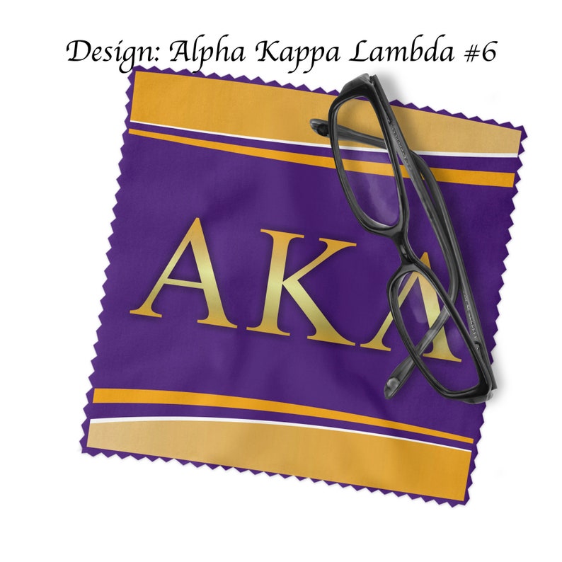 K&ouml;nnte beinhalten: Lilafarbener Mikrofaser-Reinigungstuch mit goldener Beschriftung "AKA" und goldenen Streifen. Ein Paar schwarze Brillen liegt auf dem Tuch.