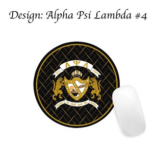May include: A black and gold round mouse mat with a Greek fraternity crest featuring a shield with a mountain, a sun, and a hand reaching out. The crest is surrounded by two golden lions and a banner that reads "&Alpha; &Psi; &Lambda; Est. 1985 Together We Shall Seek The Noblest".