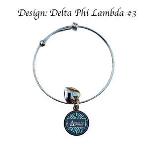 K&ouml;nnte beinhalten: Ein silbernes, ausziehbares Armband mit einem Anh&auml;nger, der ein blaues und gr&uuml;nes Wirbelmuster mit einem wei&szlig;en Dreieck und dem Text "Delta Phi Lambda" zeigt.