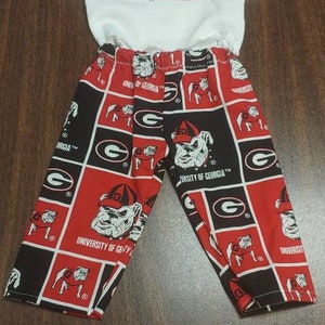 May include: A two-piece set of children's clothing featuring a white short-sleeved top and patterned pants. The pants are red, black, and white with the University of Georgia Bulldogs logo and text. The top has a small logo.