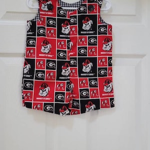 May include: Red and black sleeveless baby overall with a checkered lining and a University of Georgia bulldog pattern. The overall has buttons on the straps and a button closure in the front.