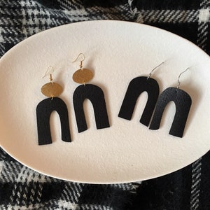 Peut inclure: Une paire de boucles d'oreilles en cuir noir avec des accents dorés. Chaque boucle d'oreille est en forme d'arche et a un petit cercle doré en haut.