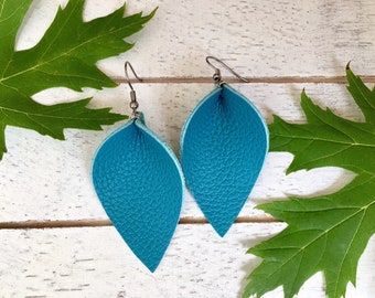 Peut inclure: Une paire de boucles d'oreilles en cuir bleu sarcelle en forme de feuilles. Les boucles d'oreilles sont suspendues à des crochets argentés.