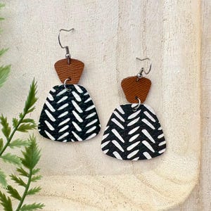 Peut inclure: Une paire de boucles d'oreilles pendantes avec un motif géométrique. Chaque boucle d'oreille comporte une petite pièce supérieure en cuir marron et une pièce inférieure plus grande à motifs noirs et blancs. Le motif est composé de lignes diagonales blanches sur fond noir.