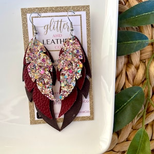 May include: Feather-shaped earrings with a layered design. The top layer is covered in glitter, with burgundy and brown leather layers below. The earrings hang from silver-coloured hooks. The earrings are displayed on a gold glitter card with the text "Glitter and Leather Co."