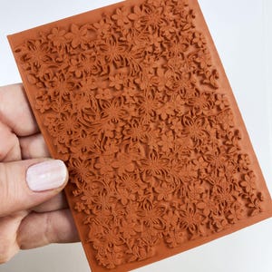 May include: A rectangular, terracotta-colored rubber stamp with a detailed floral pattern. The design features an intricate arrangement of flowers and leaves, creating a textured surface. The stamp is held by a hand with a light pink nail polish.