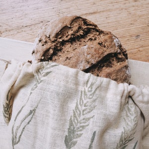 May include: A loaf of rustic brown bread in a beige linen bread bag. The bag has a green leaf pattern and a drawstring. The bread sits on a light-coloured wooden surface.
