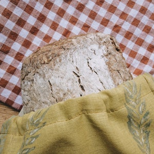 May include: A loaf of sourdough bread in a yellow fabric bag with a wheat design. The bag is sitting on a red and white gingham tablecloth.