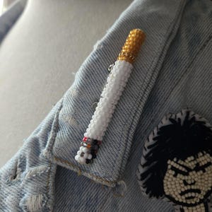 May include: A beaded cigarette brooch with a gold-coloured filter and white body, attached to a denim jacket. The brooch is made of small beads. A second, black and white beaded patch is also visible.